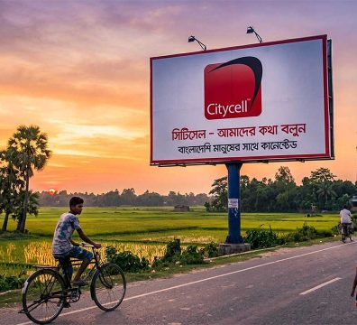 একটি বড় বিলবোর্ডে সিটিসেলের লোগো এবং বাংলা লেখা সংবলিত গ্রামীণ রাস্তার দৃশ্য যেখানে মানুষ এবং রিকশা চলাচল করছে।