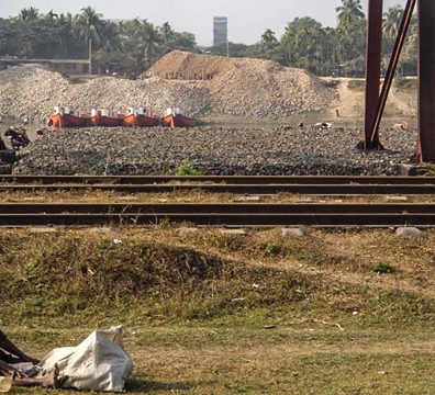 সুরমা নদীর তীরে পাথরের স্তূপের সামনে রেললাইনের পাশে বসে থাকা একজন প্রান্তিক নারী।